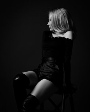 Profile silhouette of young slim pretty blonde woman in black sexy clothes and high boots sitting on stool looking aside over dark background. Fashion, style, sexy wear for women concept