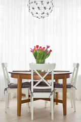 Fragment of light dining room with round wood table and bouquet or pink and red tulips in transparent glass vase on it