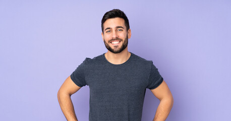 Caucasian handsome man posing with arms at hip and smiling over isolated purple background