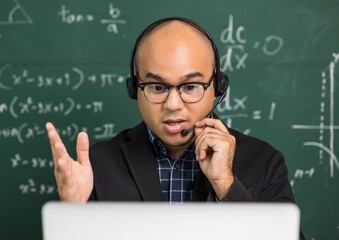 Indian young teacher man sitting wearing headset teaching online video conference live stream by laptop. Asian teacher teaching mathematics class webinar online for students learning.