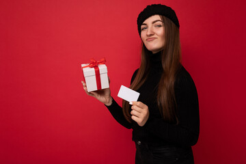 Attractive positive young brunette woman wearing black sweater and hat isolated on red background holding credit card and white gift box with red ribbon looking at camera