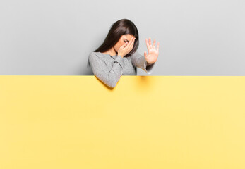 young pretty woman covering face with hand and putting other hand up front to stop camera, refusing...