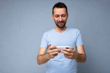 Photo shot of handsome positive good looking young man wearing casual stylish outfit poising isolated on background with empty space holding in hand and using mobile phone messaging sms looking at