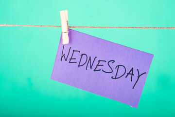  Wednesday word written on a Purple color sticky note hanging with a wire in a Cyan background.