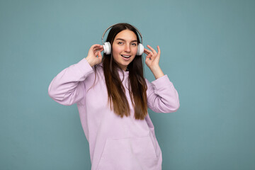 Attractive positive smiling young brunette woman wearing light purple hoodie isolated over blue background wall wearing white bluetooth headphones listening to good music and having fun looking at