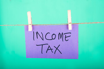 Income tax word written on a Purple color sticky note hanging with a wire in a Cyan background.