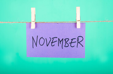  November word written on a Purple color sticky note hanging with a wire in a Cyan background.