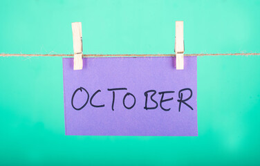  October word written on a Purple color sticky note hanging with a wire in a Cyan background.