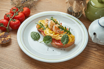 An appetizing and hearty breakfast - a sandwich with scrambled eggs, shrimps, tomatoes and hollandaise sauce, served in a white plate on a wooden background. Restaurant food