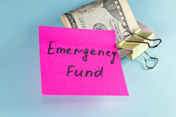 US dollars in paper clip on pastel blue background with note written EMERGENCY FUND