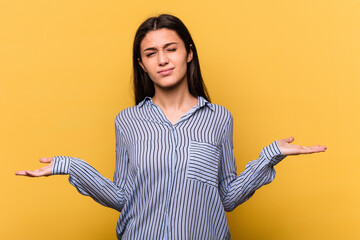 Young Indian woman isolated on yellow background doubting and shrugging shoulders in questioning...