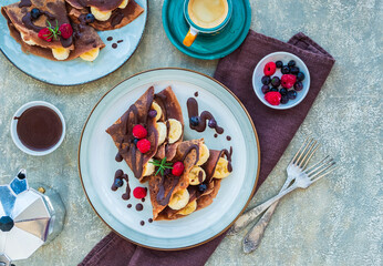 Thin chocolate pancakes stuffed with bananas, raspberries and blueberries, poured with chocolate sauce, on a gray ceramic plate on a light concrete background. Pancake recipes.