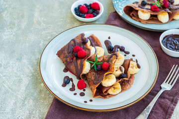 Thin chocolate pancakes stuffed with bananas, raspberries and blueberries, poured with chocolate sauce, on a gray ceramic plate on a light concrete background. Pancake recipes.