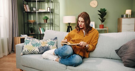 Camera approaching Caucasian young pretty happy woman resting on sofa in cozy room in house reading novel book. Female student learning at home, self-education, studying. Hobby concept