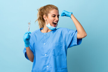 Young woman dentist holding tools over isolated blue background celebrating a victory