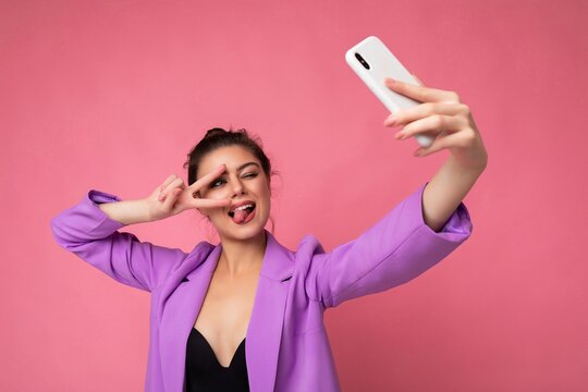 Smiling sexy woman wearing purple suit taking selfie photo on the mobile showing tongue and peace gesture phone isolated over pink background looking at cell display