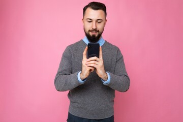 handsome good looking brunet bearded young man wearing grey sweater and blue shirt isolated on pink background with empty space holding in hand and using mobile phone communicating online looking at