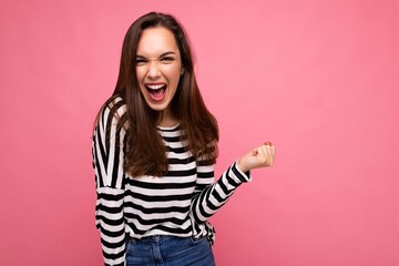 Portrait of young emotional positive happy attractive brunette woman with sincere emotions in casual striped pullover isolated on pink background with free space and celebrating winning shouting yes