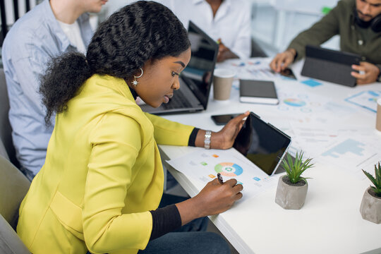 Side View Of Pretty Young Afro-american Woman In Yellow Jacket, Using Tablet And Making Notes, While Working Together At Office With Her Multiethnic Colleagues