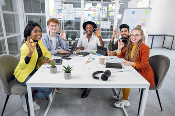 Concept of partnership and successful project. Front view of attractive positive successful diverse colleagues, sitting at the table in modern office and showing OK sign to camera