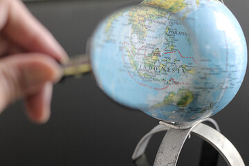 Hands holding a magnifying glass in front of a globe on a black glass table, selected focus on indonesian map.