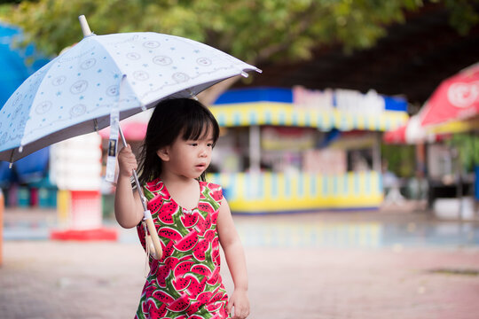 Cute Girl Walked In The Middle Of An Umbrella To Prevent The Drizzle Of Rain. Adorable Kid Wearing A Red Dress. During The Rainy Season. Kid Aged 3-4 Years Old.