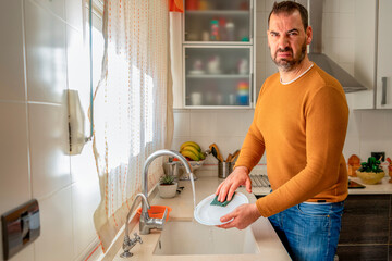 Bearded man in an orange sweater and jeans washing dishes with a jaded face and a bad attitude