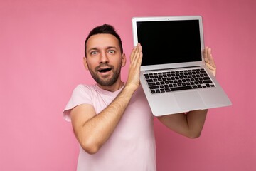 Handsome surprised and amazed man holding laptop computer looking at camera in t-shirt on isolated pink background