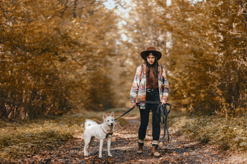 Stylish woman with backpack hiking with white dog in sunny autumn woods. Cute swiss shepherd puppy