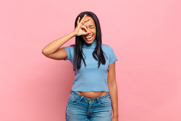 young black woman smiling happily with funny face, joking and looking through peephole, spying on...
