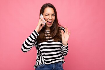 Obraz na płótnie Canvas Winking Pretty happy young woman speaking on the phone wearing striped sweater isolated over background with copy space showing ok gesture looking at camera