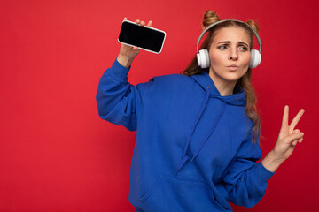 Photo of fascinating happy smiling young woman wearing stylish blue hoodie isolated over red background wearing white bluetooth wireless headphones and listening to music and showing mobile phone with