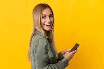 Naklejka premium Young blonde woman isolated on yellow background holding a mobile phone and with arms crossed