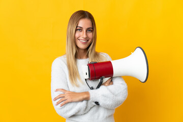 Young blonde woman isolated on yellow background holding a megaphone and smiling