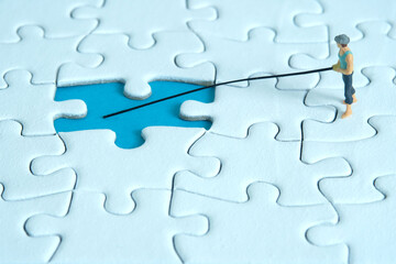 Miniature toy photography people. Fisher standing above white puzzle jigsaw piece. Isolated on blue background.