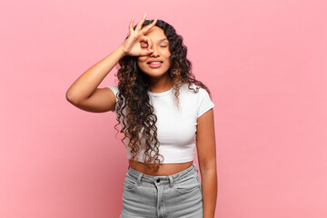 young hispanic woman smiling happily with funny face, joking and looking through peephole, spying...