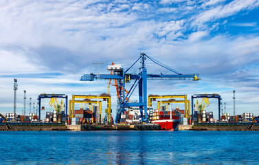 Obraz premium Sea commercial port. Loading container ship. Cranes for cargo transportation. Cargo transportation by sea. The red vessel with containers is preparing to leave the port.