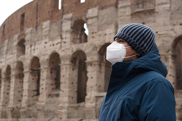 A young Italian man wearing a mask during coronavirus times in front of Colosseum, pandemic in Europe, Italy in quarantine, lockdown, ancient building