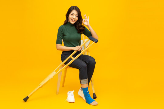 Young Asian woman sitting on chair and sprain foot using crutches and showing ok sign isolated on yellow background, Personal accident concept