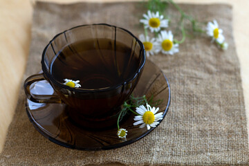 dark glass cup of Herbal tea with chamomile tea on sackcloth on a light wooden table with copy space. cup fo tea with bouquet fresh flowers and green leaves on yellow background. Healthy lifestyle. 