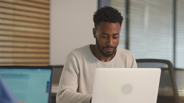 Two businessmen working at desks on laptops discussing document on screen in modern office - shot in slow motion