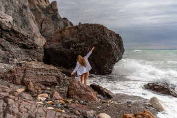 A beautiful girl in a black dress is walking on the waves, big waves with white foam. A cloudy stormy day at sea, with clouds and big waves hitting the rocks.