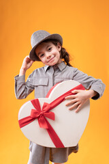 A girl touches the hat on her head and holds in her hand a wrapped gift with a bow in the shape of a large heart