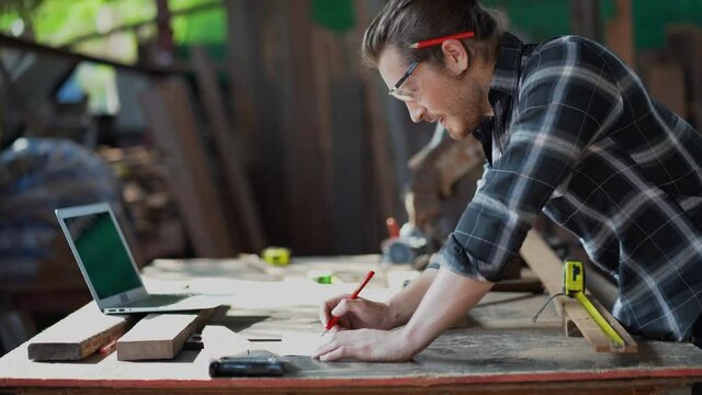 young hipster carpenter man working with computer laptop in workshop . craftsman wearing safety glasses checking order of clients or learning online