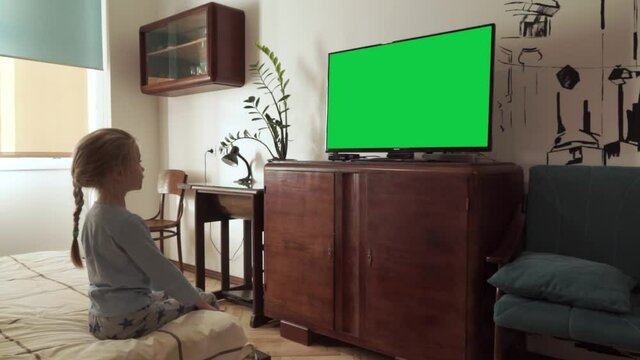 Caucasian blonde child girl watch LCD TV with green screen sitting on bed in vintage furniture decorated bedroom