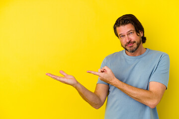 Middle age caucasian man isolated on yellow background excited holding a copy space on palm.