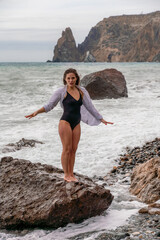 A beautiful girl in a white shirt and black swimsuit stands on a rock, big waves with white foam. A cloudy stormy day at sea, with clouds and big waves hitting the rocks.