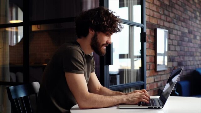 Happy young man sits down at desk, opening laptop and starts typing on keyboard of computer, side view. Smiling bearded hipster businessman working remote at home office. Guy enjoying communication.