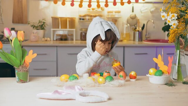 Happy Easter, kid. Girl in rabbit ears paint eggs in the kitchen of the house. Girl have fun on holiday and painting funny egg