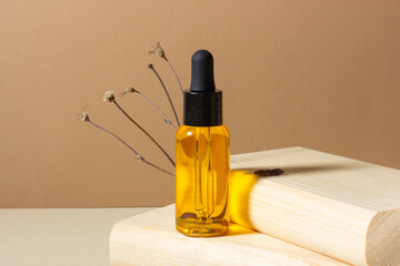 A glass cosmetic bottle with a dropper stands next to a dried flower on a beige background in bright sunlight, on a wooden podium. The concept of natural cosmetics, natural essential oil.